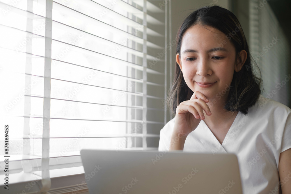 Young woman working on laptop at home, focused and engaged in her task, with natural light streaming through the blinds. Young woman working on laptop at home, focused and engaged in her task, with natural light streaming through the blinds.