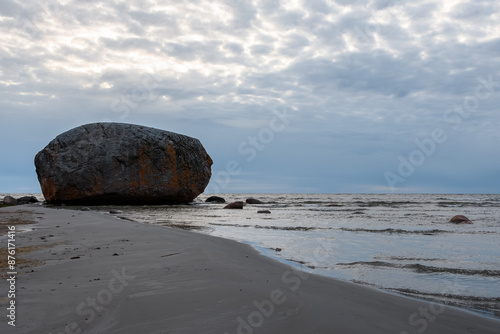 In a seascape, a large granite stone Ehalkivi (Estonia), from the Ice Age. The waters of the Baltic Sea and a wild sandy beach in the evening twilight under a beautiful gray cloudy sky, backlight