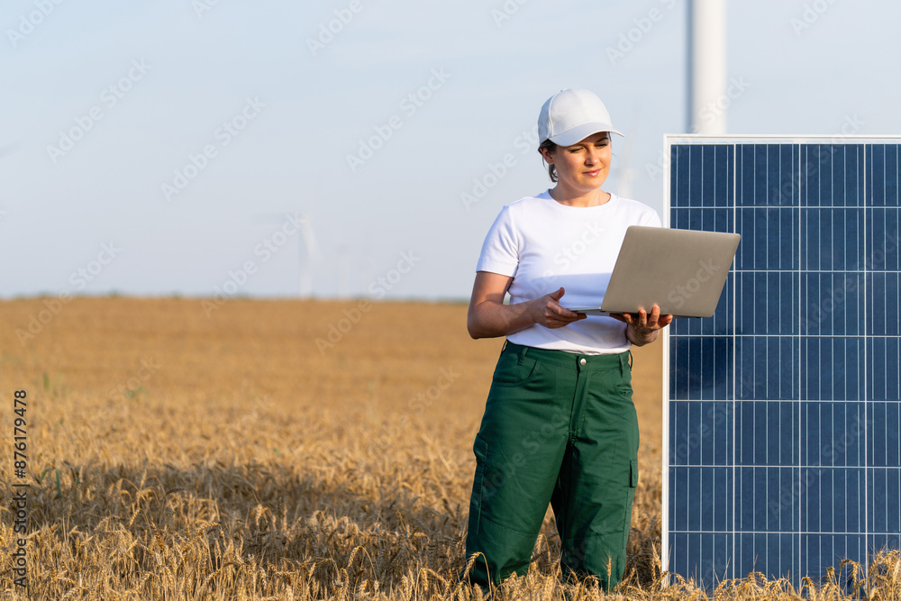 Woman farmer wearing white cap and t-shirt with laptop stands next to solar panel. Wind turbines in the background Woman farmer wearing white cap and t-shirt with laptop stands next to solar panel. Wind turbines in the background