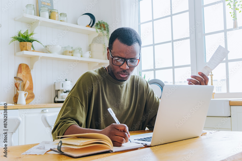 Fototapeta premium African American man writes down spending on paper checks and financial management in notebook and laptop. Budget planning, cost cutting.