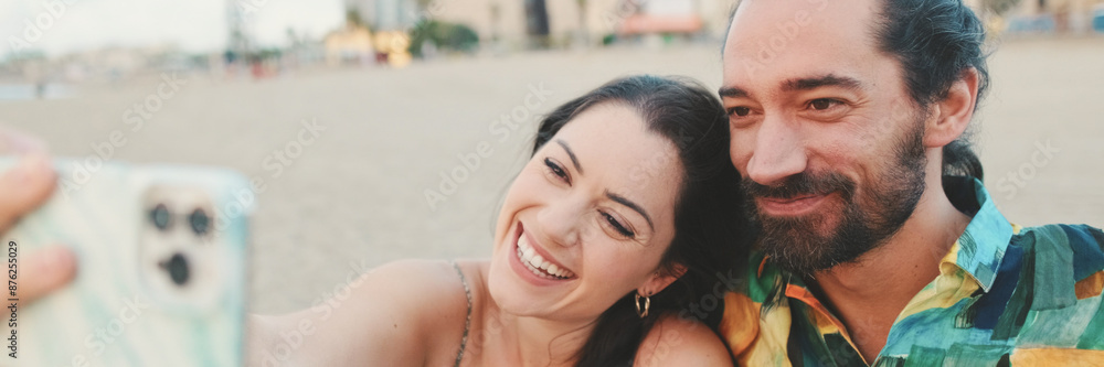 Fototapeta premium Happy couple taking selfie on mobile phone while sitting on the beach, panorama