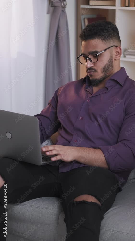 Vertical video. Distance learning. Online education. Remote study. Smart confident guy working from home using laptop on couch in modern living room interior. Vertical video. Distance learning. Online education. Remote study. Smart confident guy working from home using laptop on couch in modern living room interior.