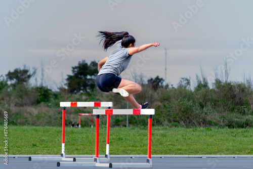 Woman practicing athletics, jumping hurdles while running. A green field in the background.