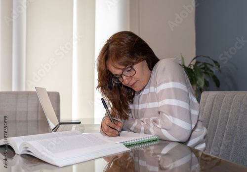 Mujer escribiendo en libreta, sentada frente a una computadora estudiando