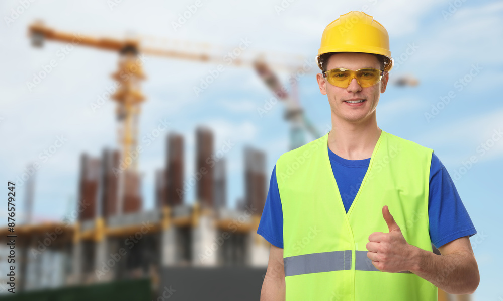 Young man wearing safety equipment and showing thumbs up at construction site. Space for text Young man wearing safety equipment and showing thumbs up at construction site. Space for text