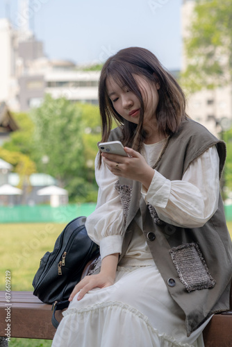 In early summer, a young Chinese exchange student in her twenties, sitting on a bench in a park near Hamamatsucho, Minato, Tokyo, operating a smartphone during the day. photo