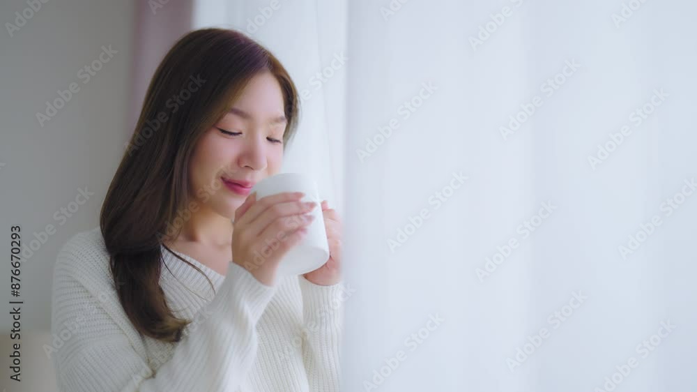 Young asian woman standing beside window and holding mug in bedroom at home, She drinking milk after wake up in the morning Young asian woman standing beside window and holding mug in bedroom at home, She drinking milk after wake up in the morning
