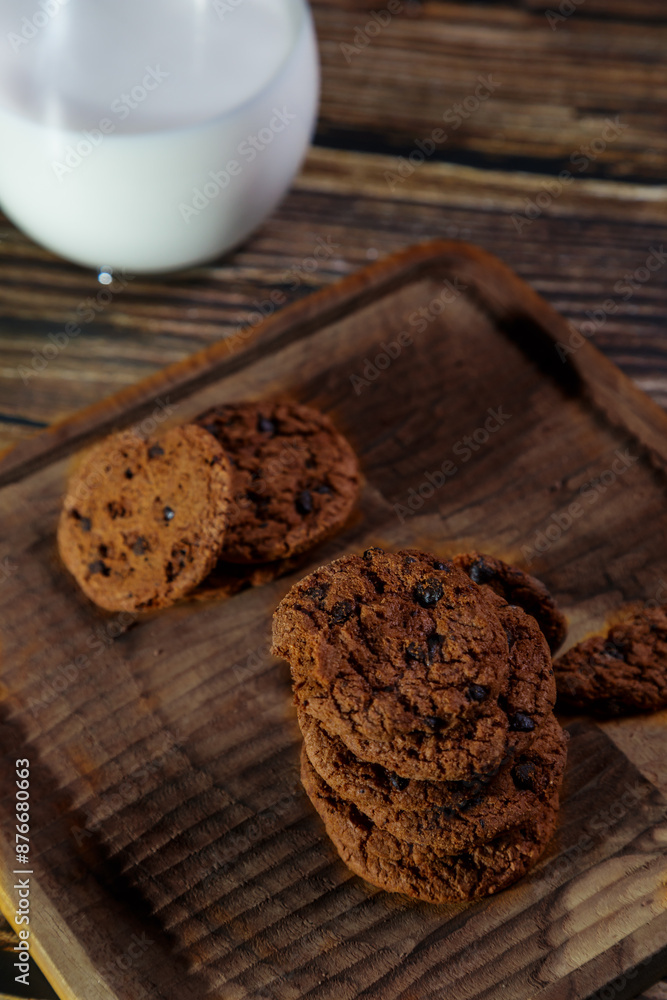 Brown chocolate chip cookies are placed on a brown wooden tray with a glass of milk and a brick wall in the background. Brown chocolate chip cookies are placed on a brown wooden tray with a glass of milk and a brick wall in the background.