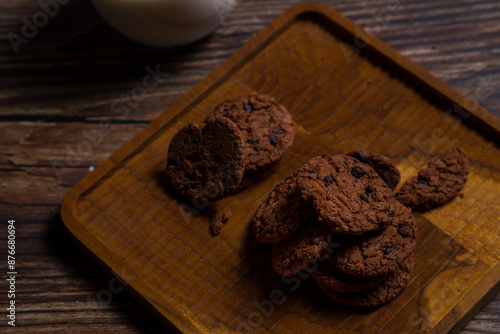 Brown chocolate chip cookies are placed on a brown wooden tray with a glass of milk and a brick wall in the background. photo