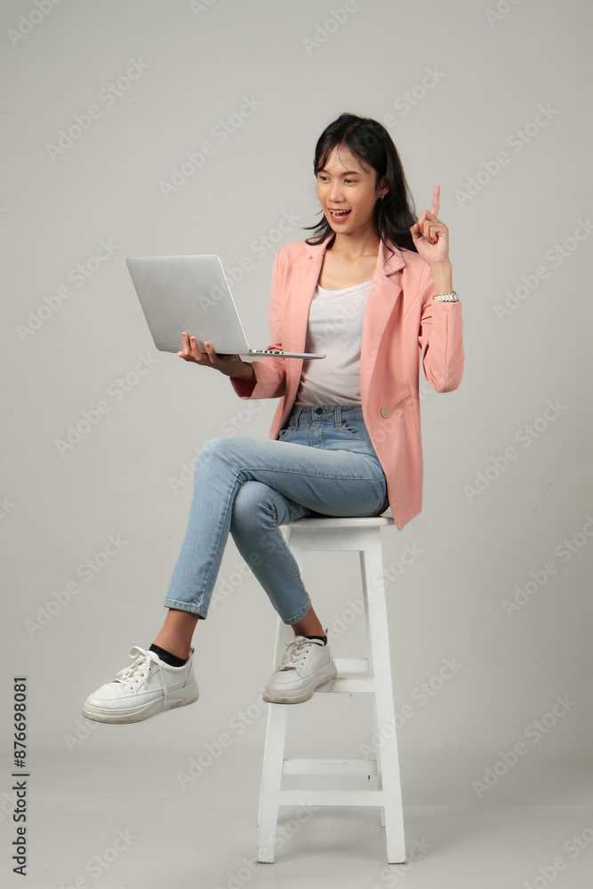 full lenght shoot of happy asian woman holding laptop computer sitting on stool with pointing hand gesture on isolated background. full lenght shoot of happy asian woman holding laptop computer sitting on stool with pointing hand gesture on isolated background.