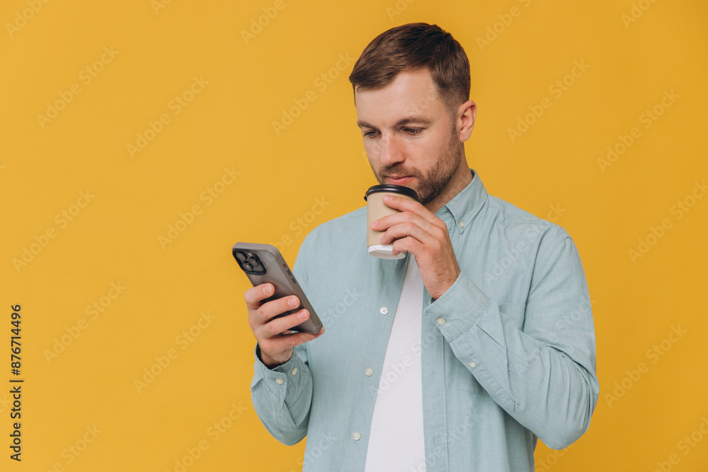 Unshaven man holding smartphone in one hand and takeaway coffee cup in another, looking at screen of phone, isolated on yellow background Unshaven man holding smartphone in one hand and takeaway coffee cup in another, looking at screen of phone, isolated on yellow background