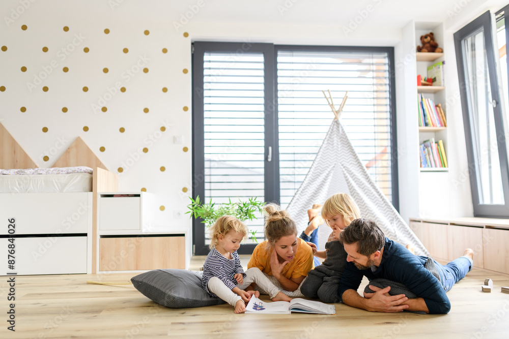 Fototapeta premium Young nuclear family playing with toys in a living room. Parents and children lying on floor, looking at children's story book, spending weekend day indoors.