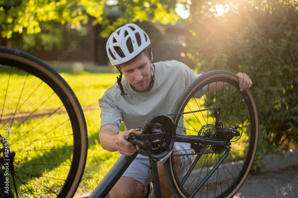 Naklejka premium man adjusts the wheel of a bicycle while standing on a paved path. Green grass and trees are in the background