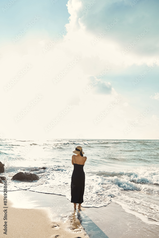 A woman in a black sundress and straw hat stands on the beach, gazing out at the ocean where gentle waves lap at the shore. A woman in a black sundress and straw hat stands on the beach, gazing out at the ocean where gentle waves lap at the shore.