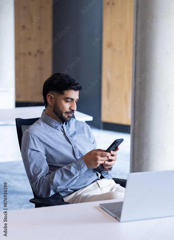Using smartphone, man sitting at desk with laptop in modern office Using smartphone, man sitting at desk with laptop in modern office