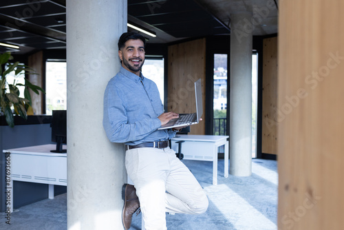 Smiling businessman leaning on pillar using laptop in modern office setting, copy space