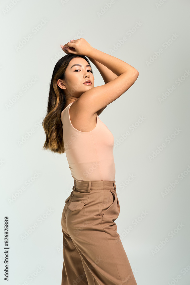 Fototapeta premium An Asian woman in a peach tank top and brown pants poses with her hand on her head.