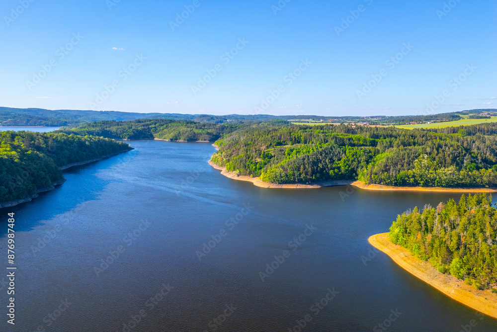 Fototapeta premium Aerial view of Orlik Water Reservoir on the Vltava River in Czechia, surrounded by forests and hills. Calm waters reflect blue sky, creating serene and idyllic landscape.