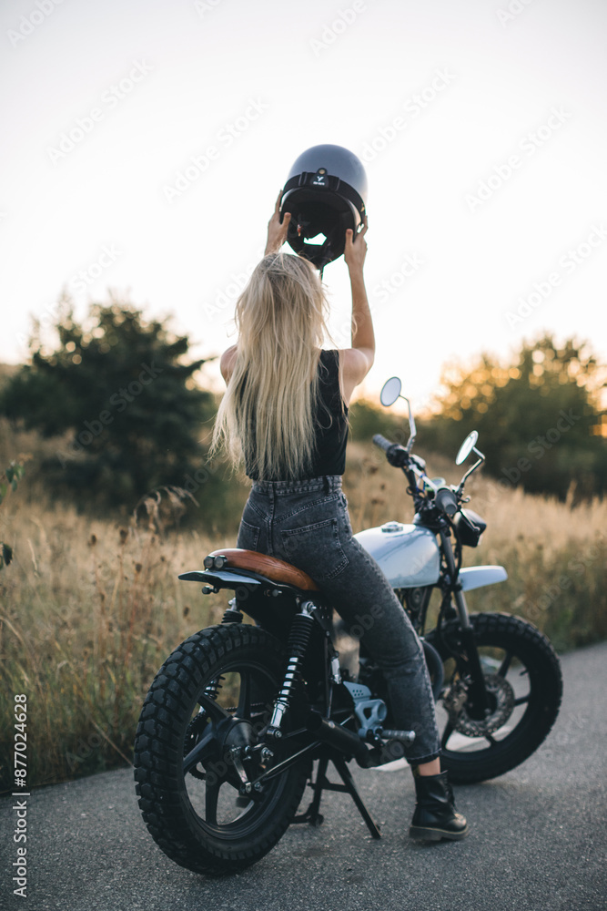 Obraz premium A cute blonde girl puts on a protective helmet while sitting on a motorcycle at sunset.