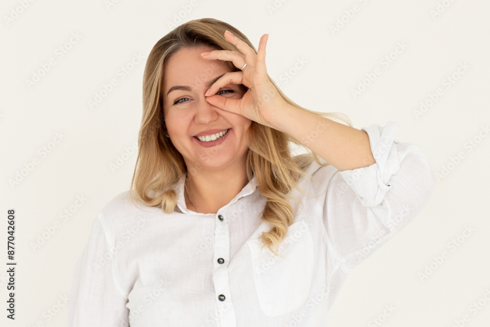 Happy young woman shows ok sign with hand on eye looking through fingers on white background. Happy young woman shows ok sign with hand on eye looking through fingers on white background.