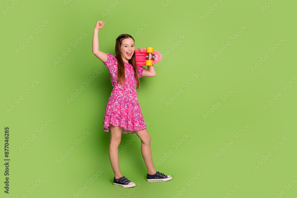 Full body portrait of lovely schoolgirl skate raise fist empty space wear pink dress isolated on green color background Full body portrait of lovely schoolgirl skate raise fist empty space wear pink dress isolated on green color background