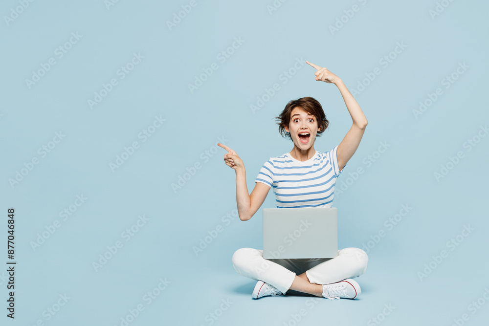 Full body shocked young IT woman wears striped t-shirt casual clothes sitting hold use work on laptop pc computer point index finger aside on area isolated on plain blue background. Lifestyle concept. Full body shocked young IT woman wears striped t-shirt casual clothes sitting hold use work on laptop pc computer point index finger aside on area isolated on plain blue background. Lifestyle concept.