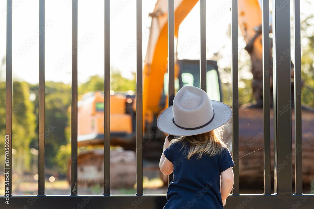 Child looking through fence at digger excavator on worksite Child looking through fence at digger excavator on worksite