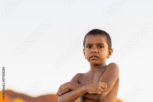 shirtless young aboriginal boy with sand on his skin against plain sky background looking at camera photo