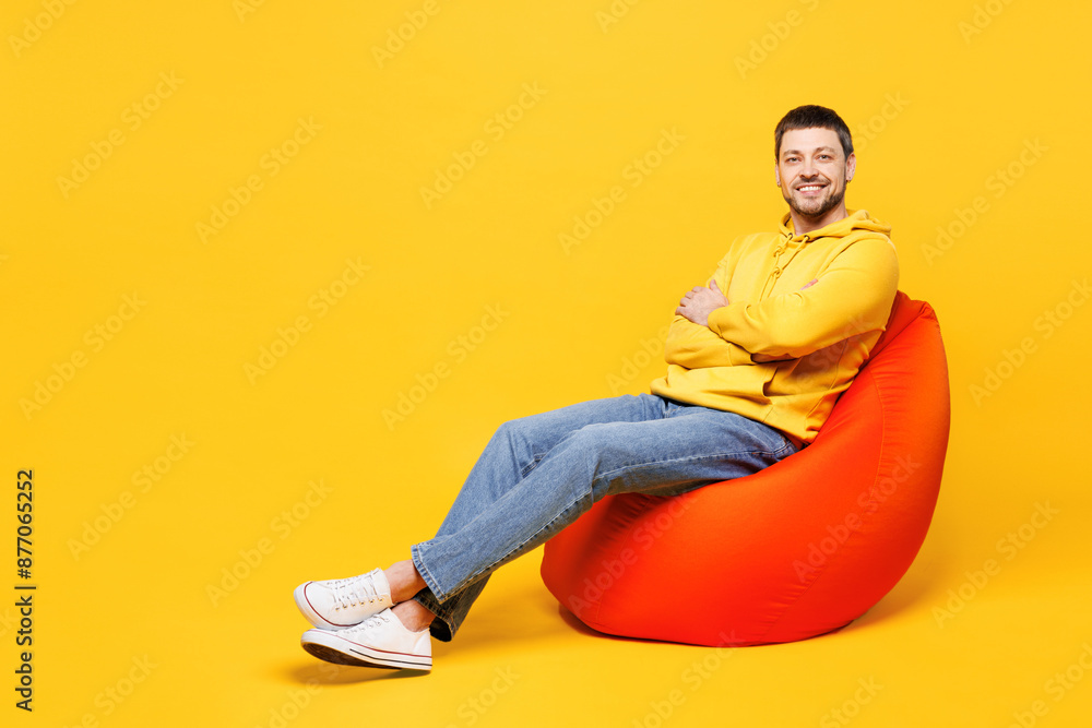 Full body young smiling happy man he wearing hoody casual clothes sit in bag chair hold hands crossed folded look camera isolated on plain yellow orange background studio portrait. Lifestyle concept. Full body young smiling happy man he wearing hoody casual clothes sit in bag chair hold hands crossed folded look camera isolated on plain yellow orange background studio portrait. Lifestyle concept.