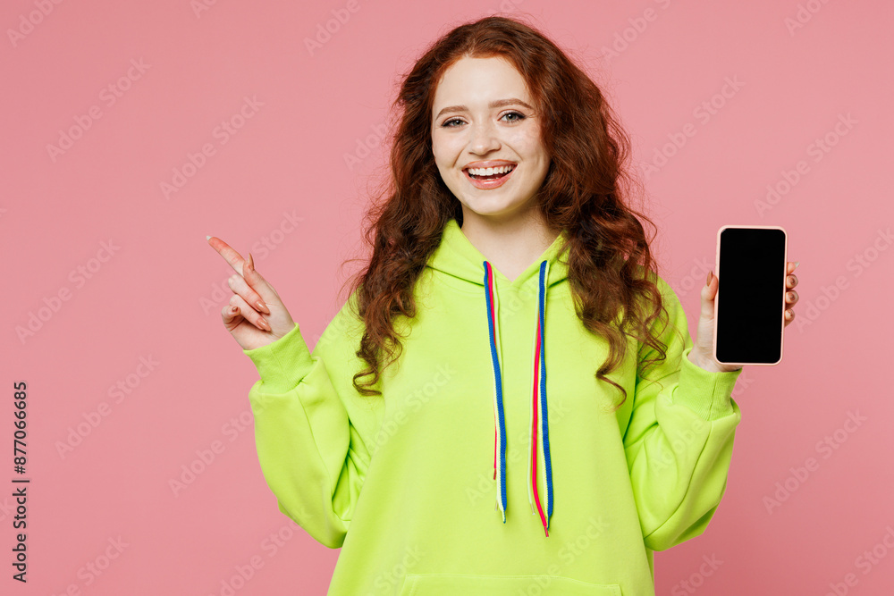 Young ginger woman wear green hoody casual clothes hold in hand use mobile cell phone with blank screen workspace area point finger aside isolated on plain pastel pink background. Lifestyle concept. Young ginger woman wear green hoody casual clothes hold in hand use mobile cell phone with blank screen workspace area point finger aside isolated on plain pastel pink background. Lifestyle concept.