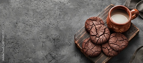 On a rustic wooden cutting board, chocolate cookies sit alongside a small clay cup of milk against a gray slate background in a horizontal view, evoking a cozy evening with copy space image. photo