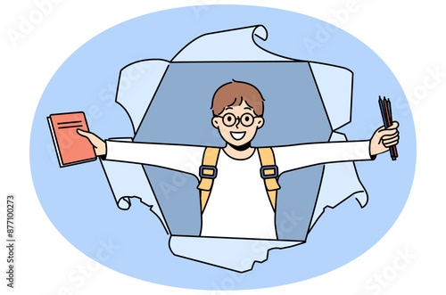 Little schoolboy with books and pencils rejoices at beginning of school year, looking out of hole in paper. Happy boy first grade student with backpack smiling for back to school concept