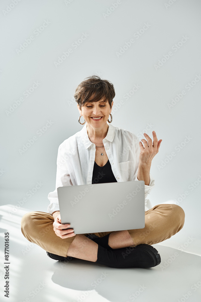 A middle-aged woman with short hair is sitting on the floor, intensely focused on her laptop in a stylish studio setting. A middle-aged woman with short hair is sitting on the floor, intensely focused on her laptop in a stylish studio setting.