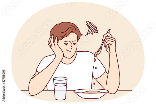 Man suffers from lack of appetite sitting at table in kitchen and does not want to eat breakfast. Depressed guy with unhappy expression melancholy looks at plate with not appetite food during diet