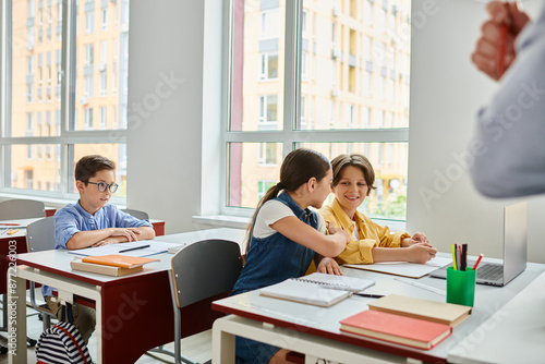 Valokuva A group of children sit at desks in a bright classroom, focusing on a man teacher giving instructions