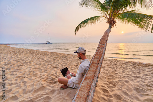 Fotografija Young nomad man freelancer sitting under a palm tree and working remotely online