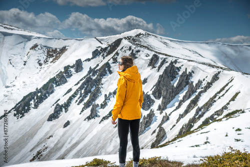 Beautiful Woman Hiking in Mountains Contemplating View of Winter Mountains. Portrait of female in orange windproof jacket feeling freedom getting to the top of the snowy mountain peak admiring scenic 