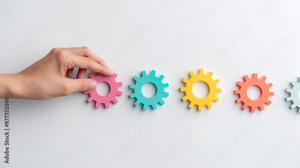 A hand placing pink, blue, yellow, and orange gears in a row against a white background. A hand placing pink, blue, yellow, and orange gears in a row against a white background.