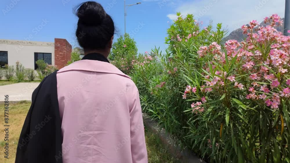 Woman from her back walking by flowers and greenery with a building and blue sky in the background Woman from her back walking by flowers and greenery with a building and blue sky in the background