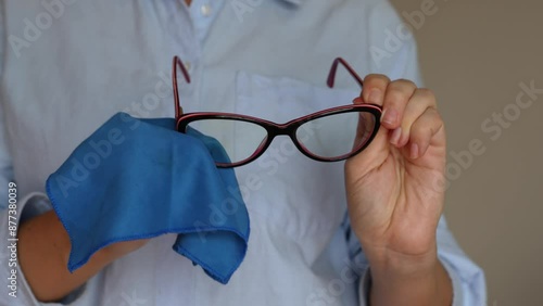 Cropped shot of a young woman wiping her glasses with a blue cloth on beige background. Fashionable glasses for working at a computer with a blue filter lenses. Anti blue light and rays photo