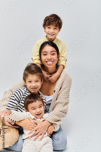 A young Asian mother lovingly poses with her children in a studio, set against a soft grey background.