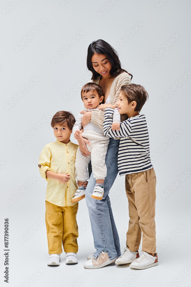 A young Asian mother tenderly holding her children, stand close by in a studio setting against a grey background. A young Asian mother tenderly holding her children, stand close by in a studio setting against a grey background.