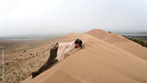 A man hikes up the Singing Dunes in the Altyn-Emel National Park, Kazakhstan. The vast desert landscape stretches behind him, with rolling dunes and distant mountains shrouded in mist. 