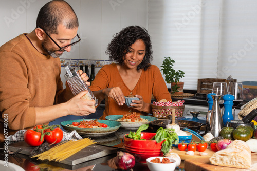 Young Hispanic couple in the kitchen preparing food. Man grates cheese while woman puts seasoning on delicious pasta