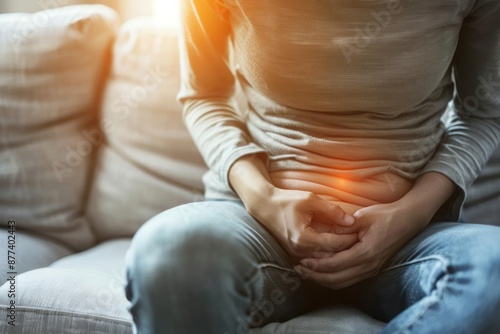 Person clutching their abdomen in pain while sitting on a couch, with a look of discomfort on their face, abdominal pain associated with colitis photo