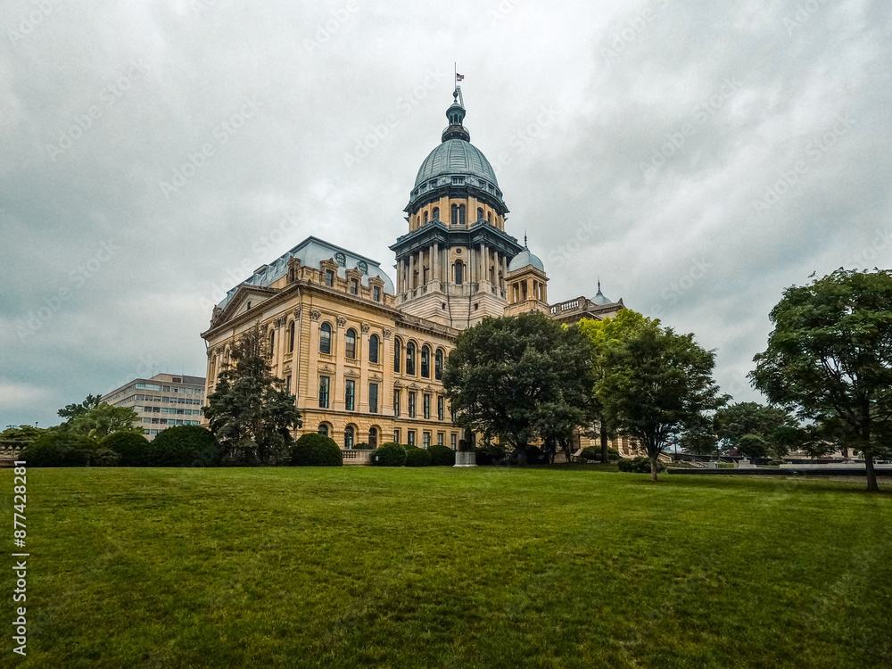 Obraz premium Long view of the Illinois State Capitol Building in Springfield, IL, USA. Cloudy overcast skies overhead. Springtime scene with a lush green lawn and full trees.