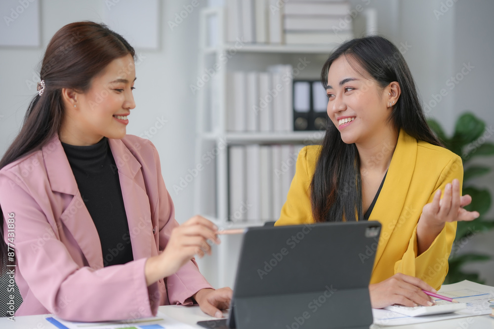 Two young businesswomen are sitting at their desk in the office, smiling and discussing while working on a new project using a tablet Two young businesswomen are sitting at their desk in the office, smiling and discussing while working on a new project using a tablet