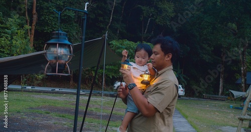 A father holding his child Enjoying Camping Trip in Forest with illuminated by lanterns at dusk, The scene captures a serene and bonding moment in nature.