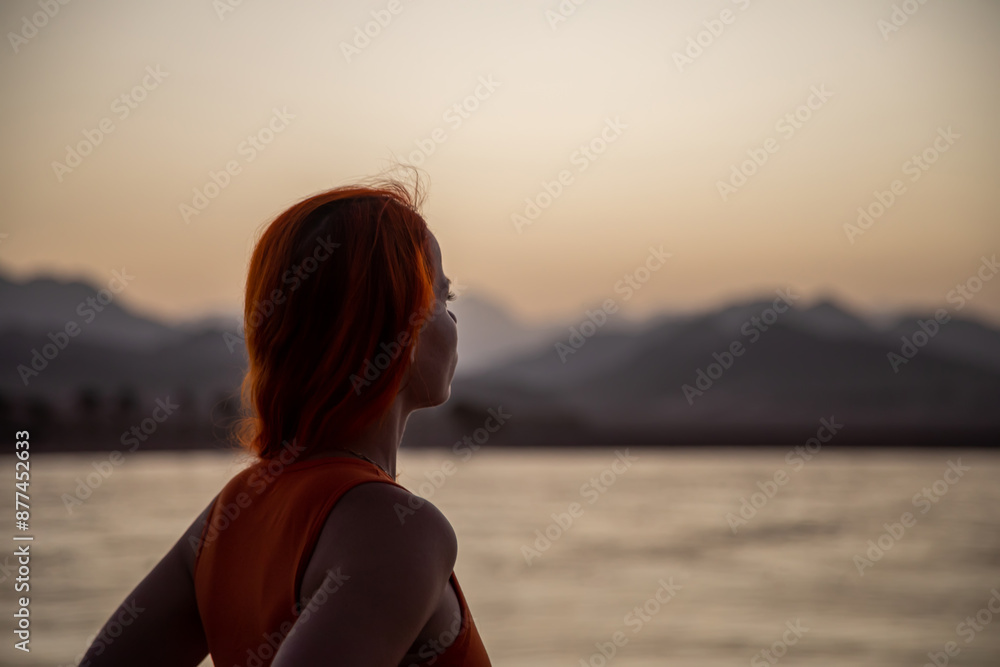 Beautiful girl stands at sunset by the sea. Sunny summer evening. Girl with red hair. Beautiful girl stands at sunset by the sea. Sunny summer evening. Girl with red hair.