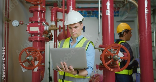 Engineer in safety gear inspecting red industrial pipes while holding using laptop in a factory environment, ensuring proper maintenance and functionality.